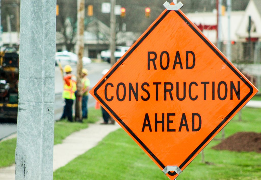 Street. Construction workers in the background. Construction sign in the foreground that reads: "Road Construction Ahead."