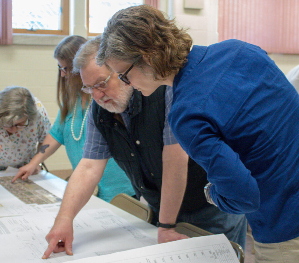 Three women, a man, stand over behind a table, pointing at maps.